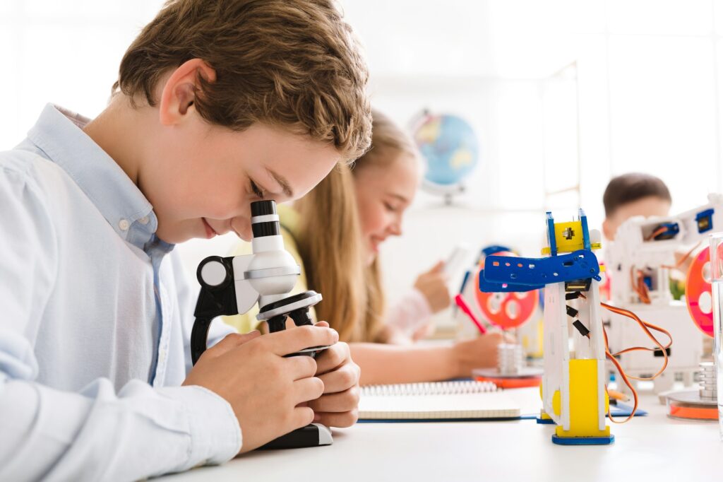 Smart schoolboy looking at microscope with robot nearby
