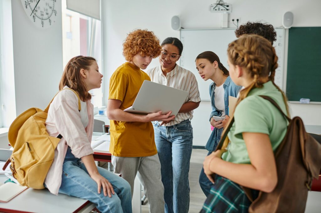 redhead schoolboy showing laptop to african american teacher near classmates, back to school concept