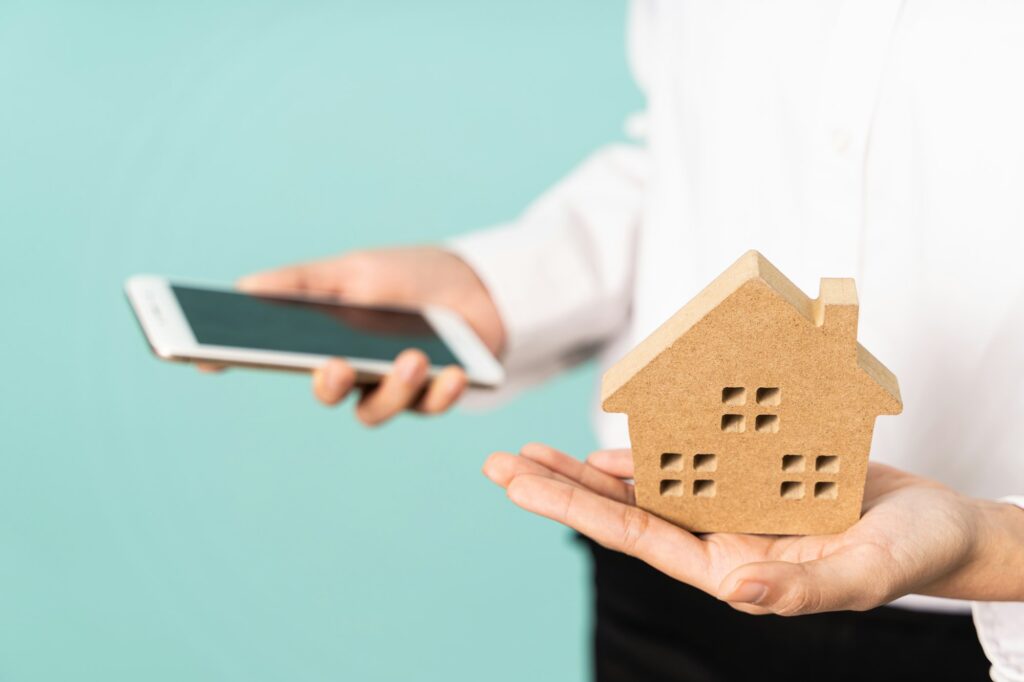 Closeup of hands holding house model and mobile on blue background. Real estate and housing concept.